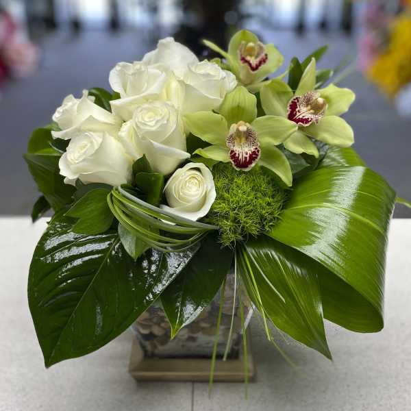 White roses and green orchids in a square glass vase with broad leaves