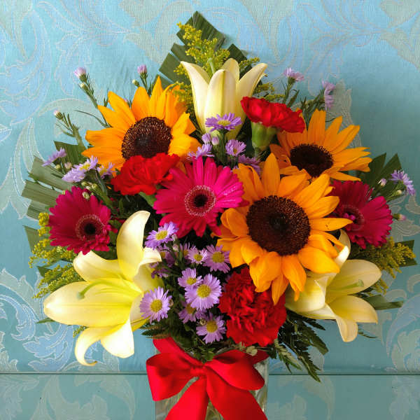 Mixed bouquet with sunflowers, lilies, gerbera daisies, and a red ribbon in a glass vase