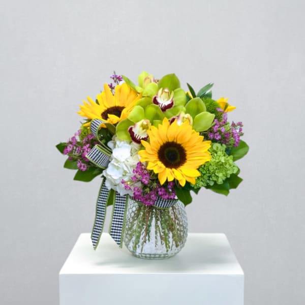 Bright arrangement of yellow sunflowers, green orchids, and mixed blooms in a clear glass vase with checked ribbon.