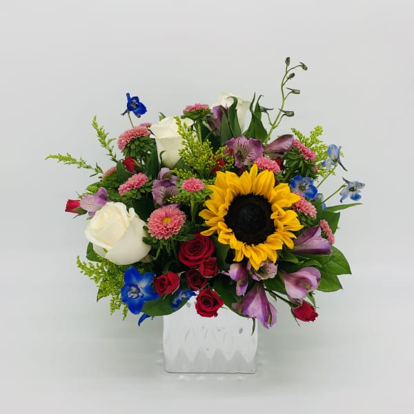 Colorful mixed bouquet with sunflower, roses, mums, and blue blooms in a white glass cube vase
