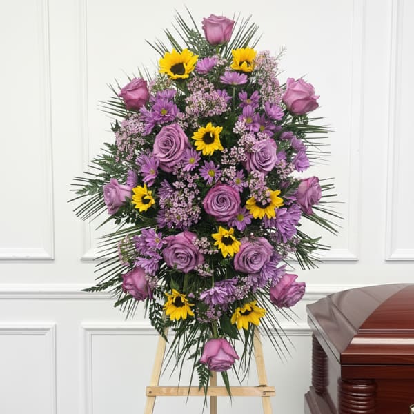 Tall standing spray of lavender roses and yellow daisies on an easel beside a wooden casket.