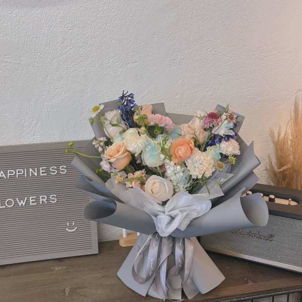 Pastel bouquet of roses, carnations, and blue flowers wrapped in gray paper