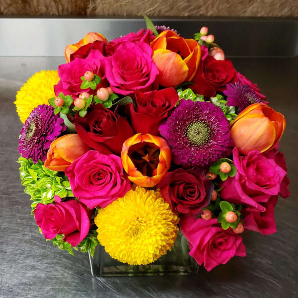 Bright mixed bouquet of roses, tulips, and mums in a square glass vase
