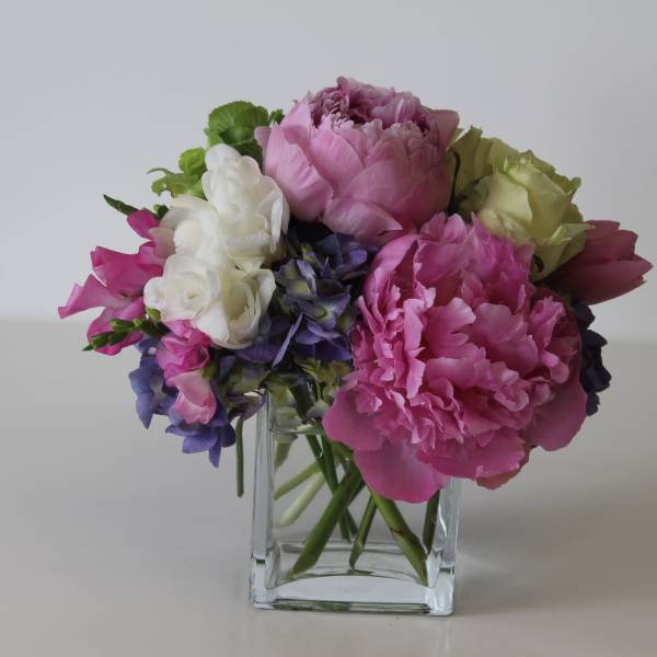 Low arrangement of pink peonies, white and green roses, and purple blooms in a clear glass cube vase