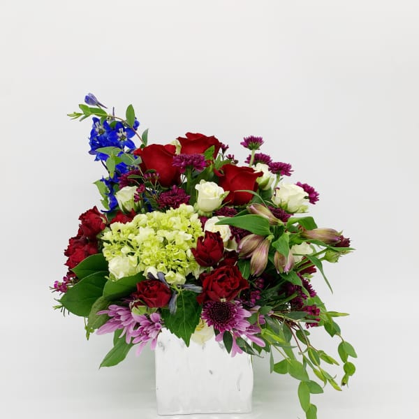 Mixed red, white, green, and purple flower arrangement in a white square vase with trailing foliage
