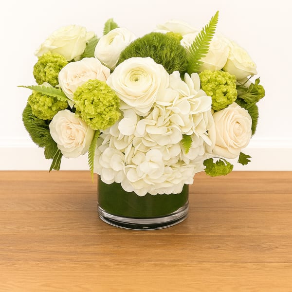Low arrangement of white hydrangeas and cream roses with green accents in a clear glass cylinder vase