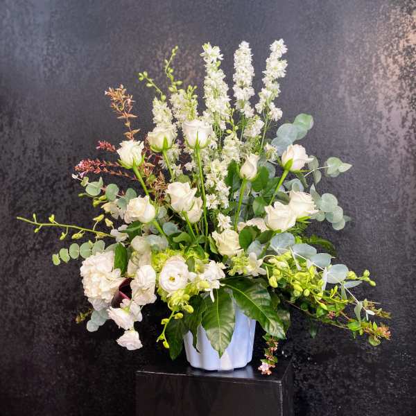 Tall white and green floral arrangement with roses and orchids in a white pot against a dark background