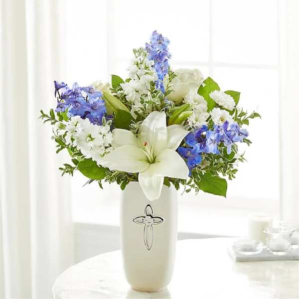 White lilies, roses, and blue delphinium in a tall white vase with a silver cross.