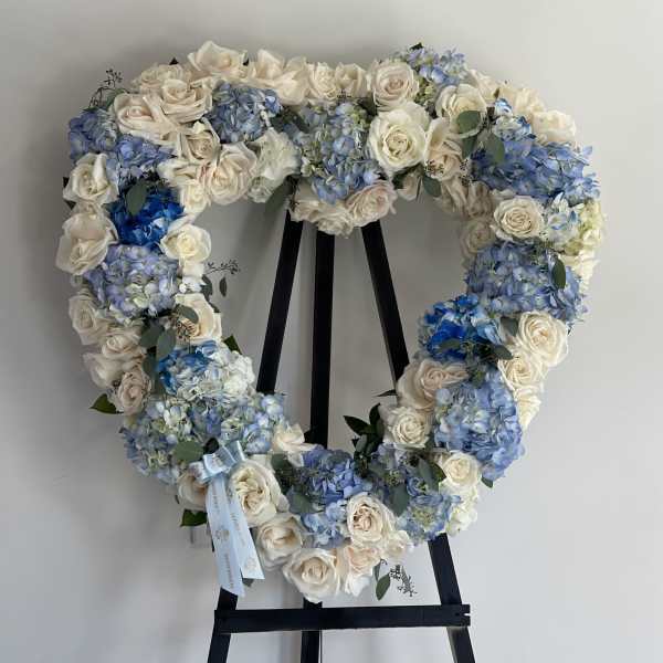 Heart-shaped floral wreath with white roses and blue hydrangeas on a black stand