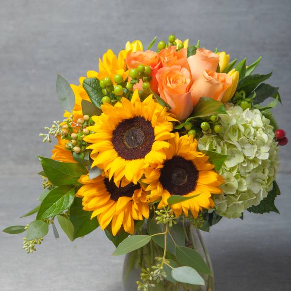 Bouquet of sunflowers, peach roses, and pale green hydrangea in a glass vase