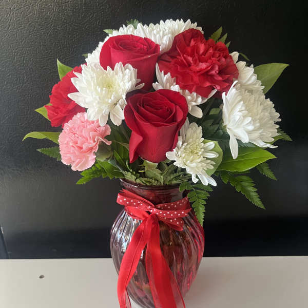 Red roses, white daisies, and pink carnations in a glass vase with a red ribbon