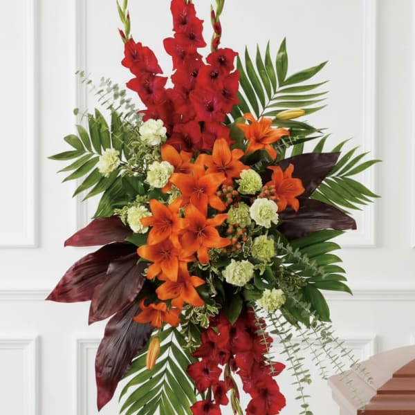 Tall standing spray of red gladiolus, orange lilies, and green carnations on an easel beside a casket.