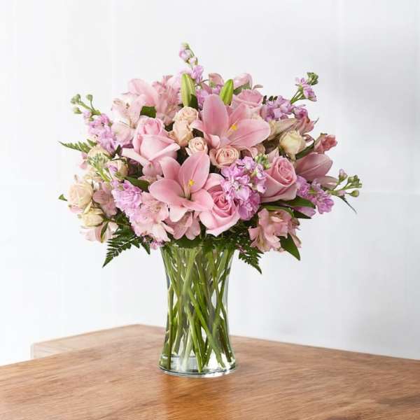 Pink lilies, roses, and mixed flowers arranged in a clear glass vase on a wooden table
