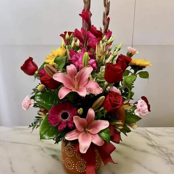 Tall mixed arrangement with pink lilies, red roses, yellow blooms in an orange patterned vase with ribbon