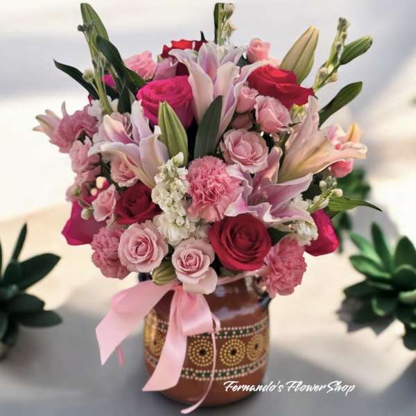 Pink and red bouquet with lilies, roses, and carnations in a painted ceramic vase with a pink bow.