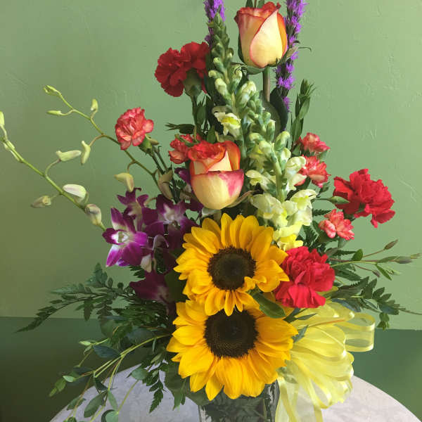 Tall mixed bouquet with sunflowers, roses, orchids, and carnations in a glass vase
