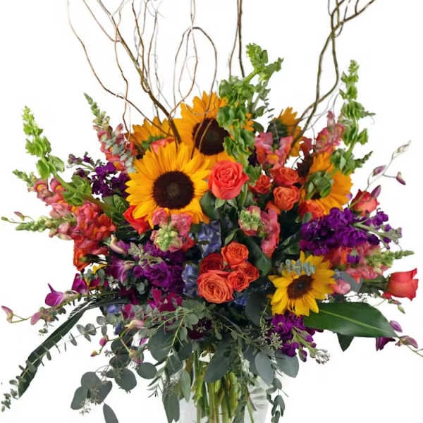 Tall mixed arrangement with sunflowers, orange roses, purple flowers, and curly branches in a clear glass vase