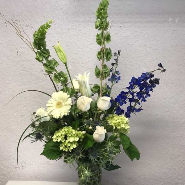 Tall floral arrangement with white roses, lilies, and blue flowers in a glass vase
