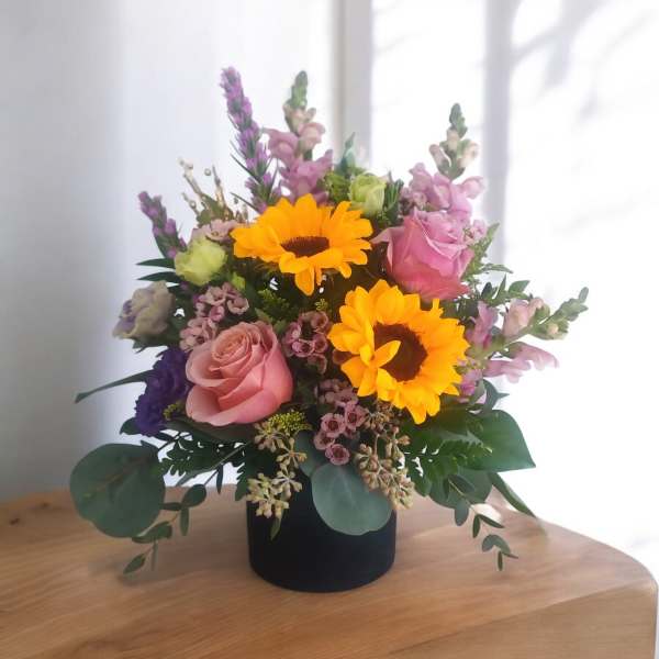 Bright arrangement with yellow sunflowers, pink roses, and purple blooms in a black vase.