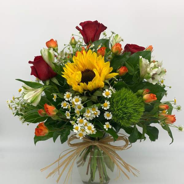 Mixed bouquet with a sunflower, red and orange roses, daisies, and white blooms in a glass vase with raffia bow.