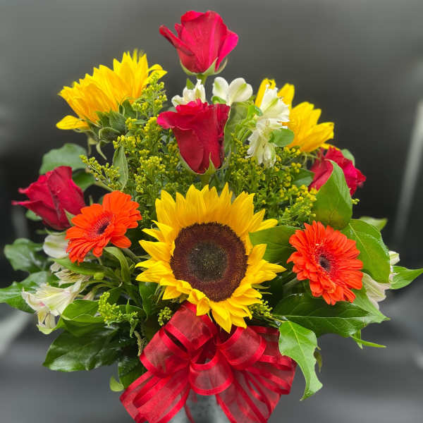 Bright mixed arrangement with sunflowers, red roses, orange gerberas and white blooms in a glass vase with red bow