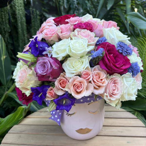 Mixed roses and purple flowers in a face-shaped vase with a polka-dot ribbon