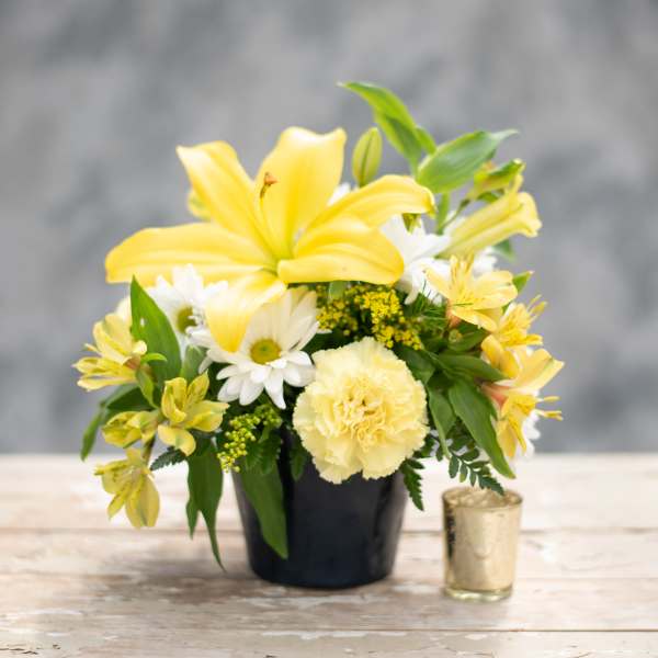 Yellow lilies and white daisies in a black pot with a small candle beside it