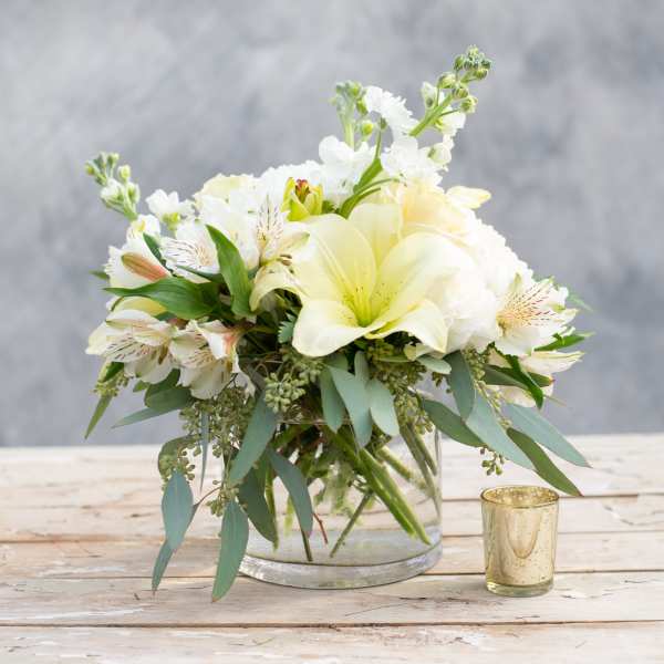 White and pale yellow bouquet in a clear glass vase beside a gold votive
