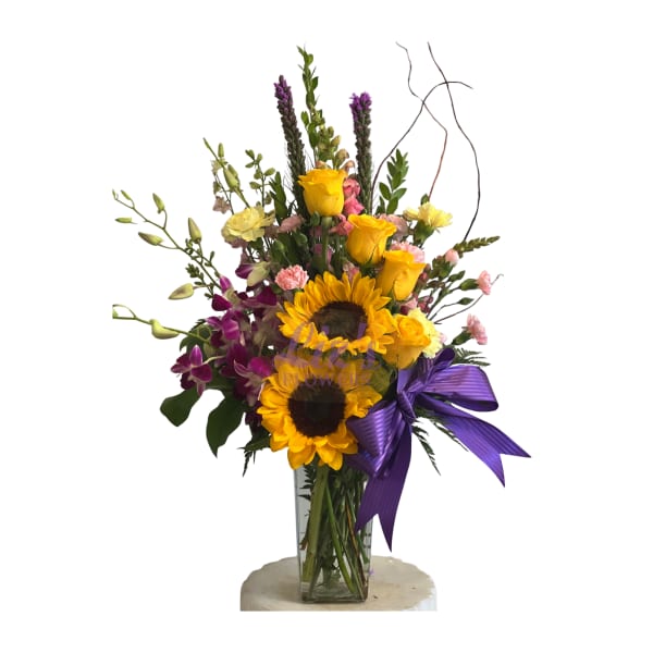 Tall arrangement with sunflowers, yellow roses, pink carnations and purple orchids in a glass vase with a purple bow.