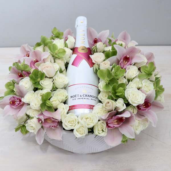 Bottle of champagne nestled in a low ring of white roses, green blooms, and pink orchids in a white bowl