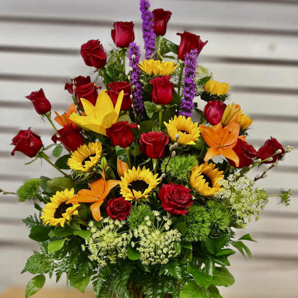 Tall mixed arrangement of red roses, sunflowers, lilies, and purple accents in a clear glass vase