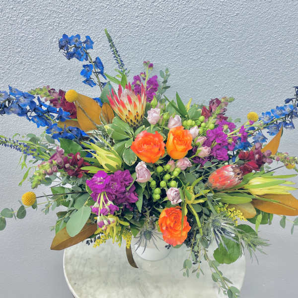 Bright mixed bouquet with orange roses, blue flowers, and pink blooms in a white vase
