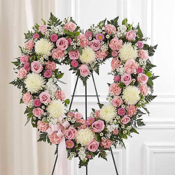 Heart-shaped standing wreath of pink roses, carnations, and white mums with a bow on a metal easel