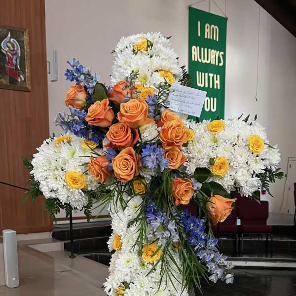 Large floral cross on an easel with white chrysanthemums, orange roses, and blue flowers