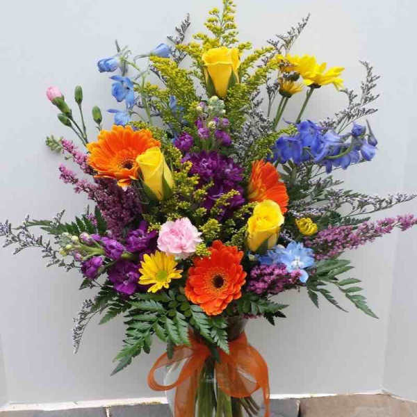 Tall mixed arrangement of bright yellow roses, orange gerberas, and purple and blue flowers in a glass vase