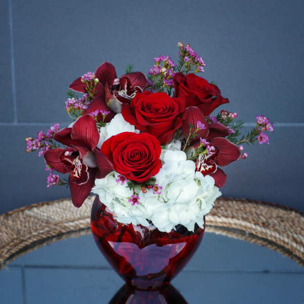 Red roses, burgundy orchids, and white hydrangeas in a red glass vase