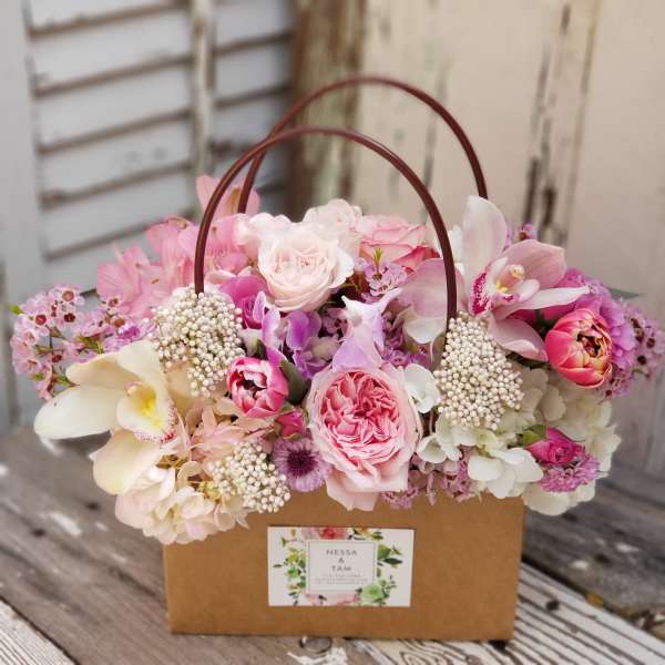 Pink and white roses, orchids, and mixed blooms arranged in a kraft paper tote with handles.