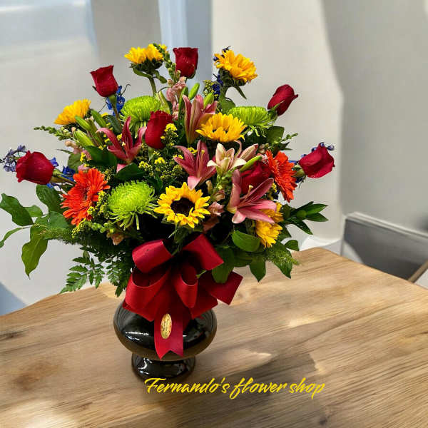 Mixed bouquet of red roses, yellow blooms, and pink lilies in a black vase with a red bow