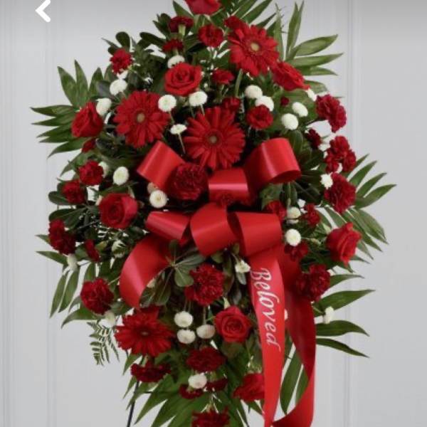Tall red and white funeral spray on an easel with roses, daisies, and a red 'Beloved' ribbon