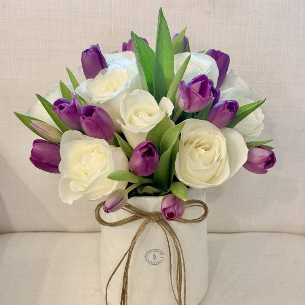 White roses and purple tulips arranged in a white vase with a twine bow.