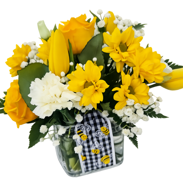 Yellow roses, daisies, and tulips with white carnations in a clear cube vase with a bee ribbon