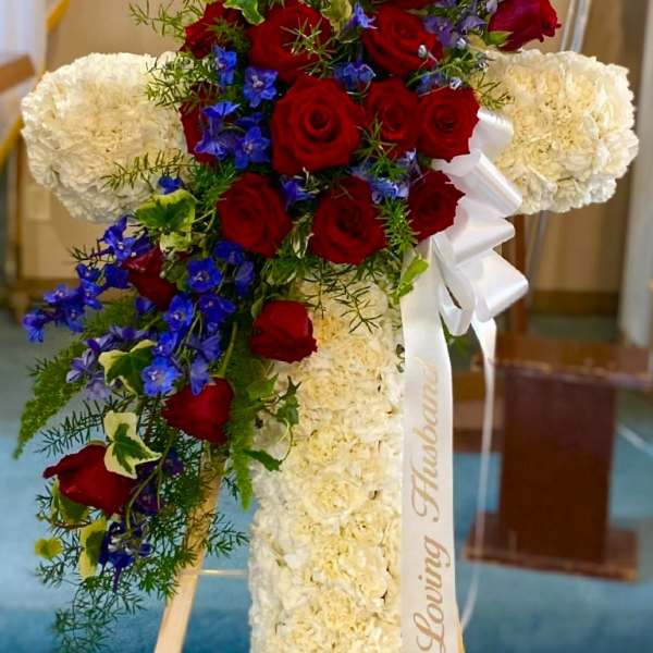 Cross-shaped floral tribute with red roses, white blooms, and blue flowers on an easel