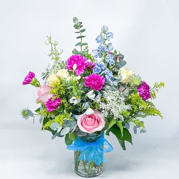 Mixed bouquet of pink, blue, and white flowers in a glass vase with a blue ribbon