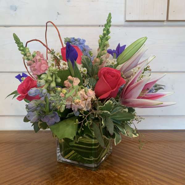 Mixed bouquet of pink roses, lilies, and blue flowers in a square glass vase