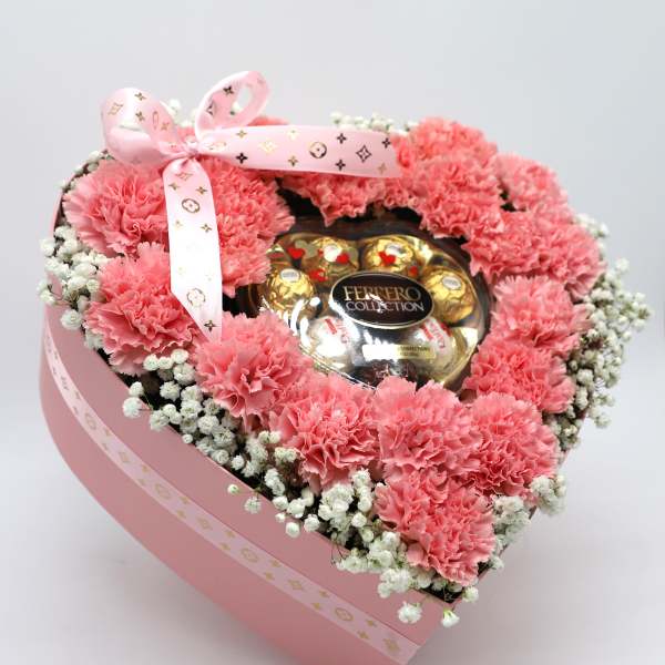 Pink carnations arranged around a Ferrero Rocher chocolate box in a pink heart-shaped box