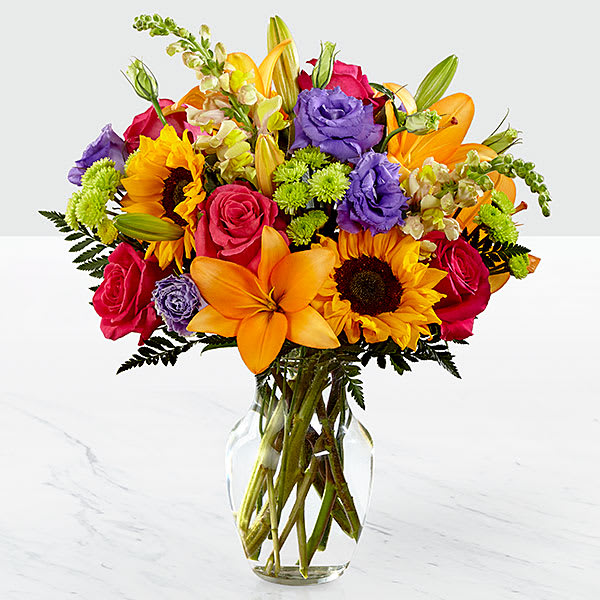 Colorful bouquet of roses, lilies, sunflowers, and purple blooms in a glass vase