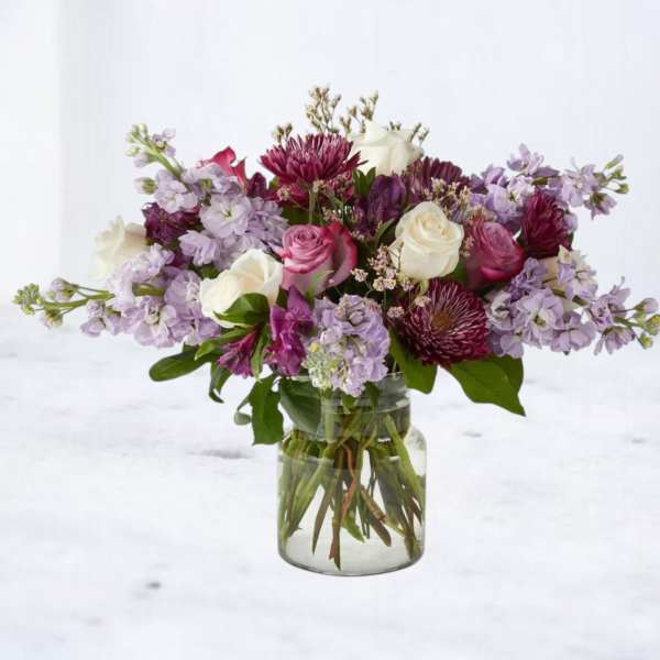 Mixed bouquet of purple, pink, and white flowers arranged in a clear glass jar vase
