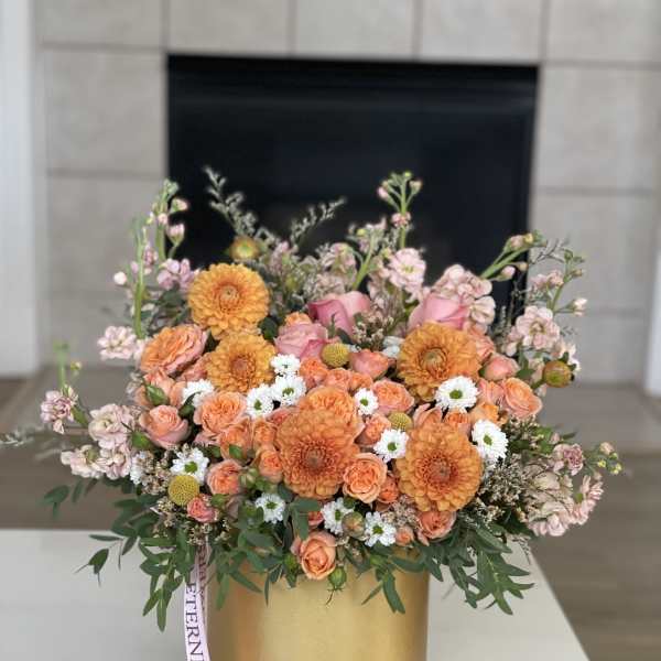 Orange and pink floral arrangement in a gold box with a ribbon