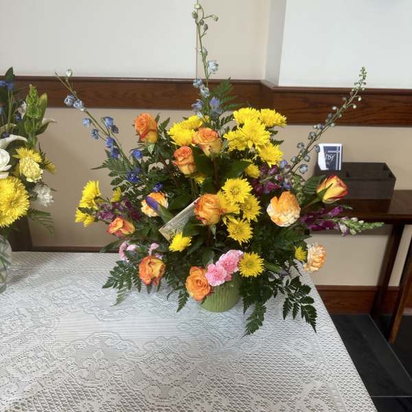 Tall mixed arrangement of yellow mums, orange roses, and blue delphinium in a green vase on a lace tablecloth