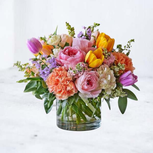 Low glass vase of pink peonies, tulips, orange carnations, and mixed pastel blooms.